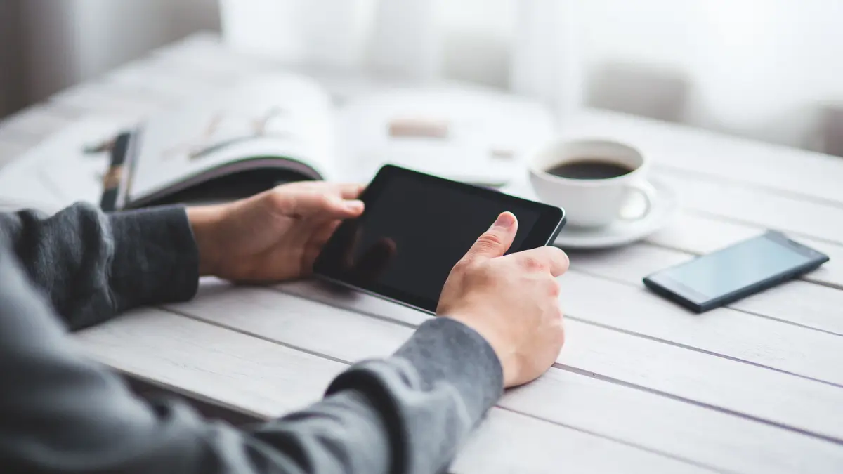 Mains tenant une tablette sur une table, près d'un téléphone et d'une tasse