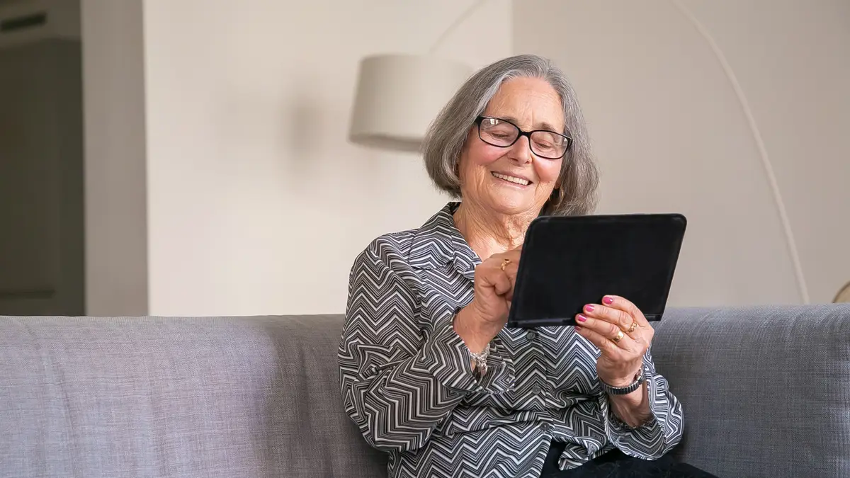 Femme âgée souriante, une tablette dans les mains, assise dans un canapé